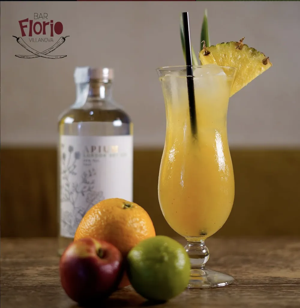 Un cocktail giallo con fetta di ananas in un bicchiere da hurricane, accanto a una bottiglia di gin Apium e a frutta fresca