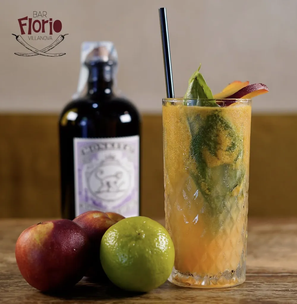 Un cocktail giallo con basilico e fetta di pesca in un bicchiere alto lavorato, accanto a una bottiglia di gin e frutta fresca su un tavolo di legno