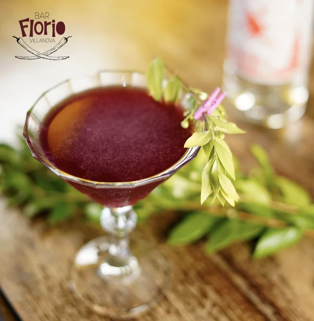 Un cocktail rosso rubino in una coppa da martini con un rametto di erbe fresche come guarnizione, su un tavolo di legno