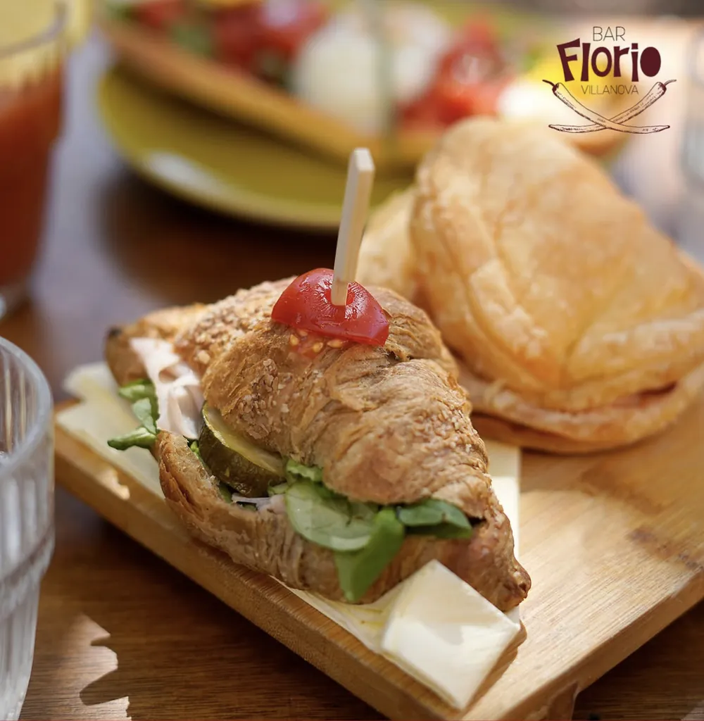 Un croissant salato farcito con prosciutto, formaggio, zucchine grigliate e insalata su un tagliere di legno, con una sfoglia accanto