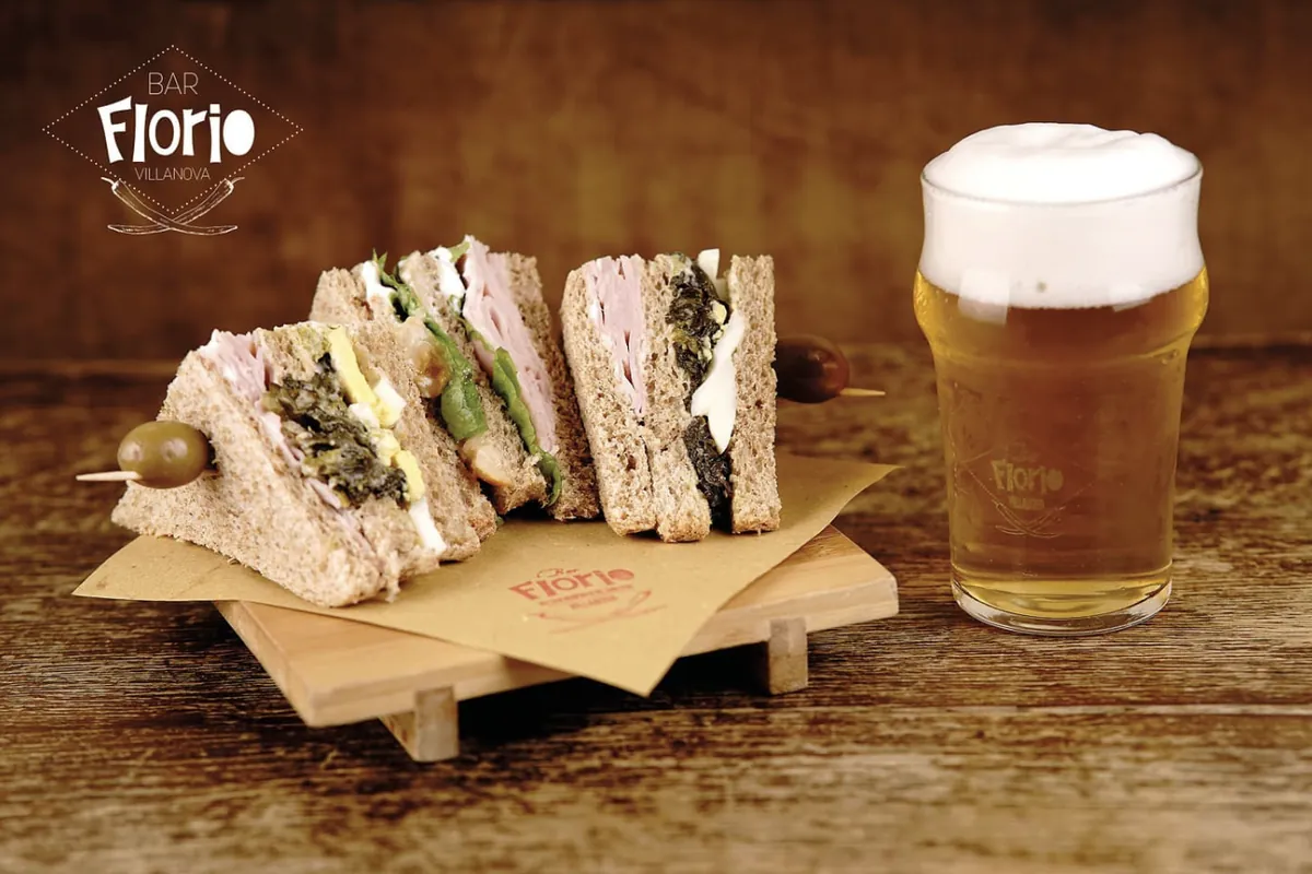 Tre tramezzini di pane integrale farciti, infilzati con olive e pomodorini, accanto a un bicchiere di birra chiara con il marchio del Bar Florio